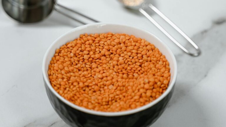 Red lentils in a bowl on marble, surrounded by measuring spoons.